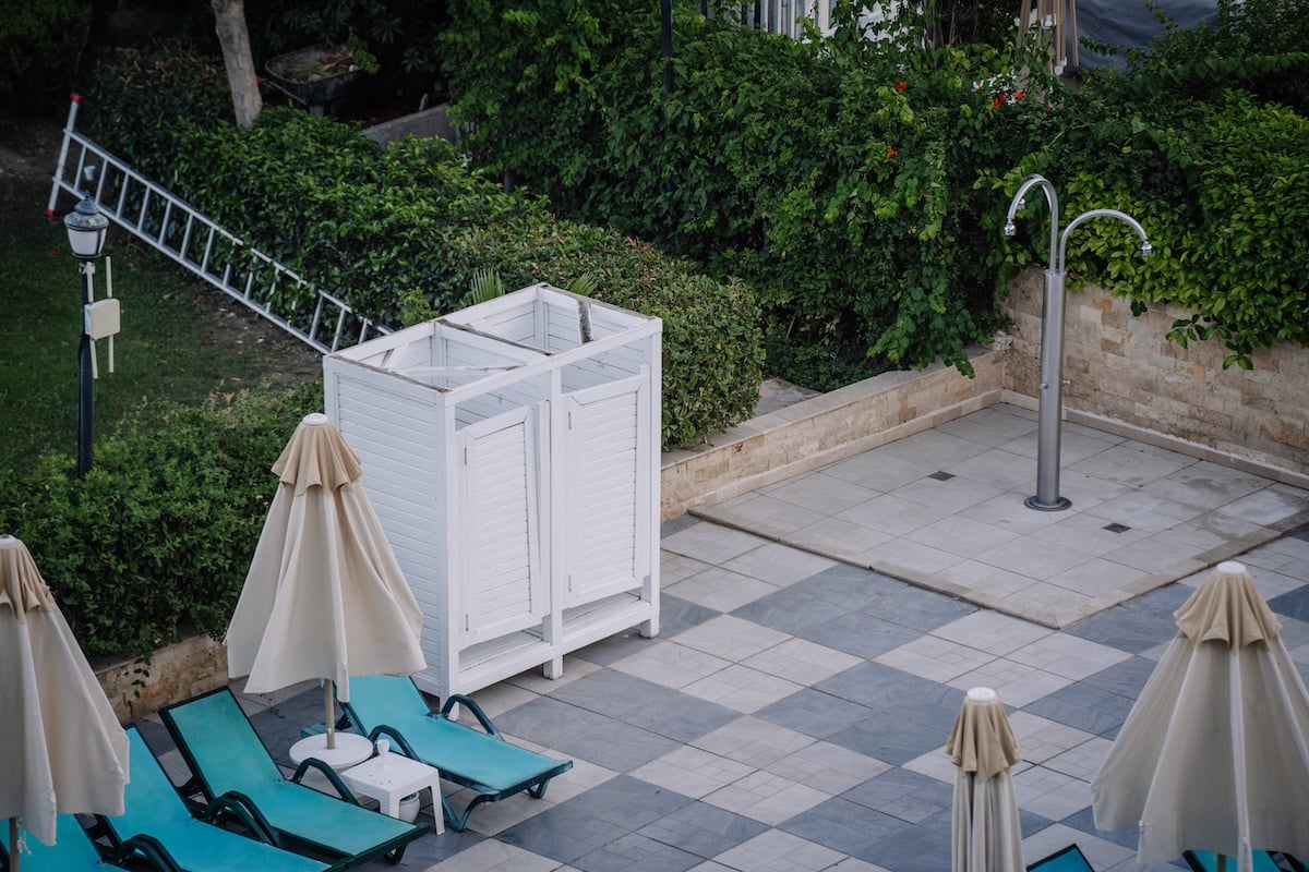 An outdoor shower area beside a white changing cabin and sun loungers with beige umbrellas, located in a landscaped poolside space with tile flooring and greenery.