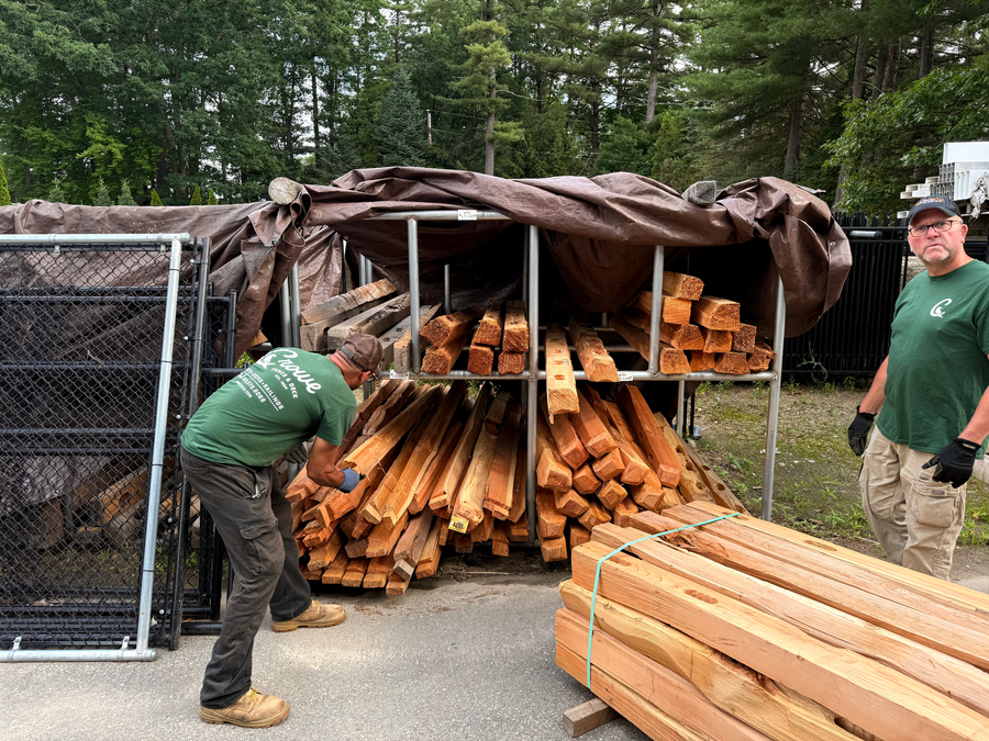 Crowe Fence team workers in green shirts unloading cedar lumber from an outdoor storage rack
