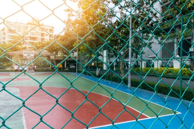 A fence encloses the basketball court where a group of school children are playing basketball