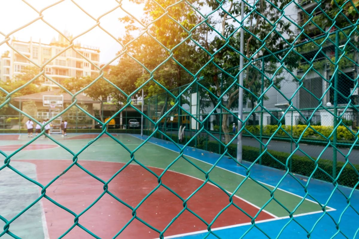 A fence encloses the basketball court where a group of school children are playing basketball