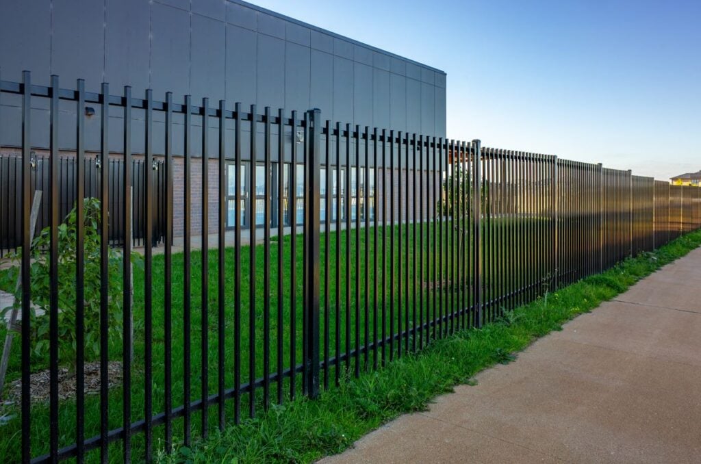 A black vertical metal security fence lines a path in front of a contemporary institutional or school building