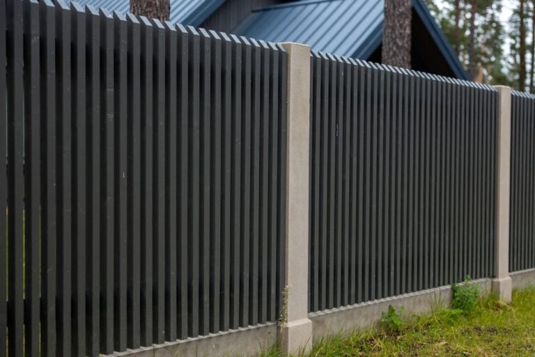 Modern dark wooden fence stands along concrete posts beside pine trees and residential building, providing perimeter security