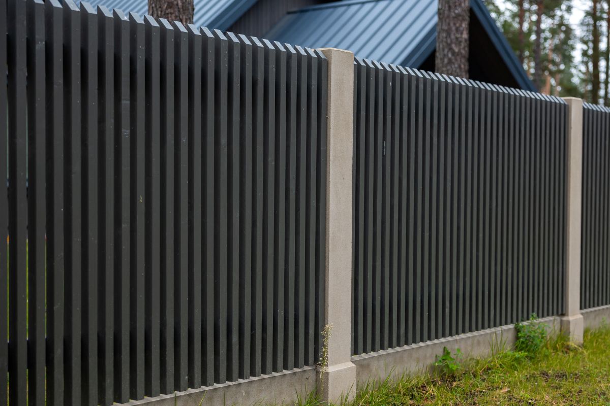 Modern dark wooden fence stands along concrete posts beside pine trees and residential building, providing perimeter security